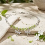 Pulsera Libélula Plata 925 con cristales verdes y dos dijes de libélulas, presentada sobre piedra natural con lino y flores blancas.