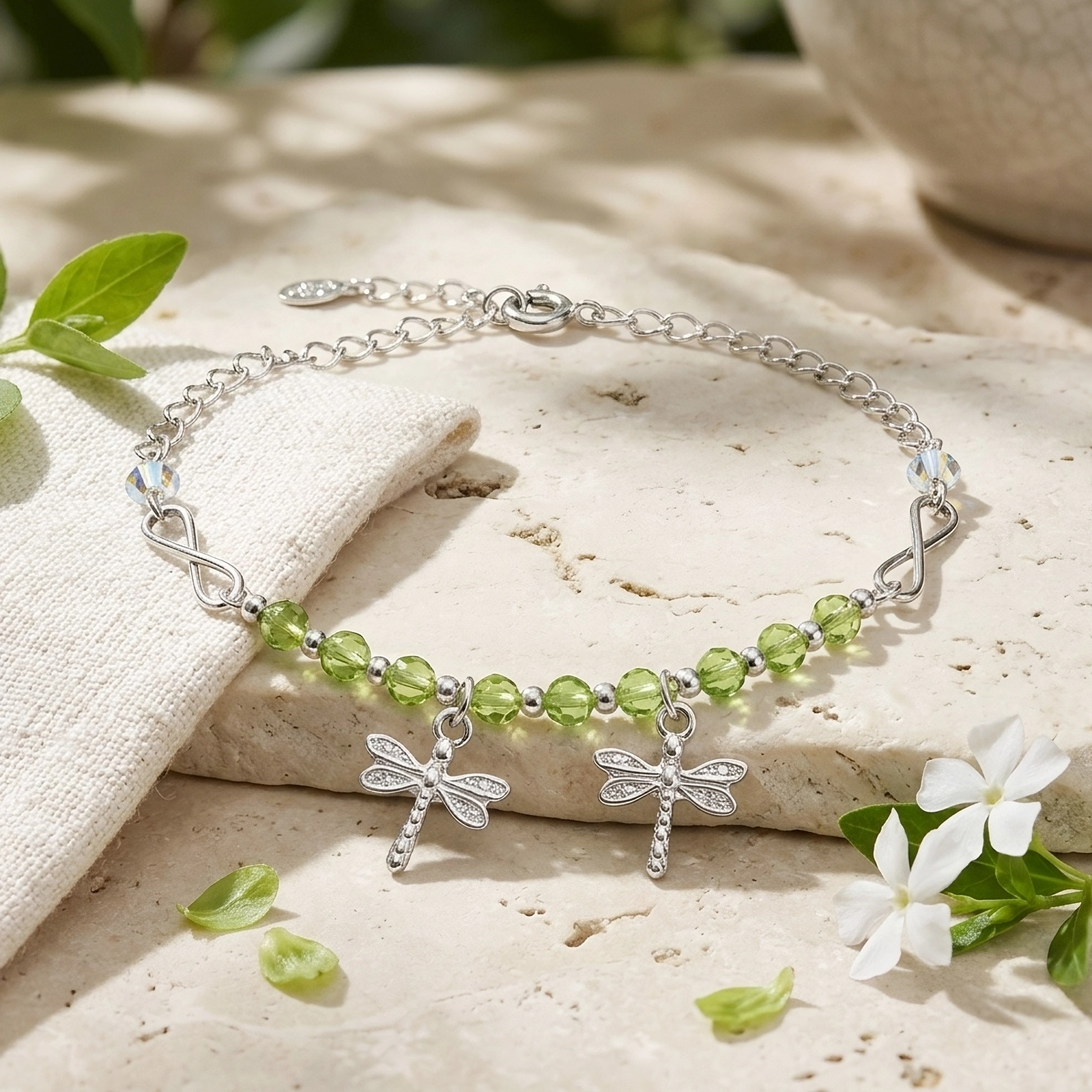 Pulsera Libélula Plata 925 con cristales verdes y dos dijes de libélulas, presentada sobre piedra natural con lino y flores blancas.