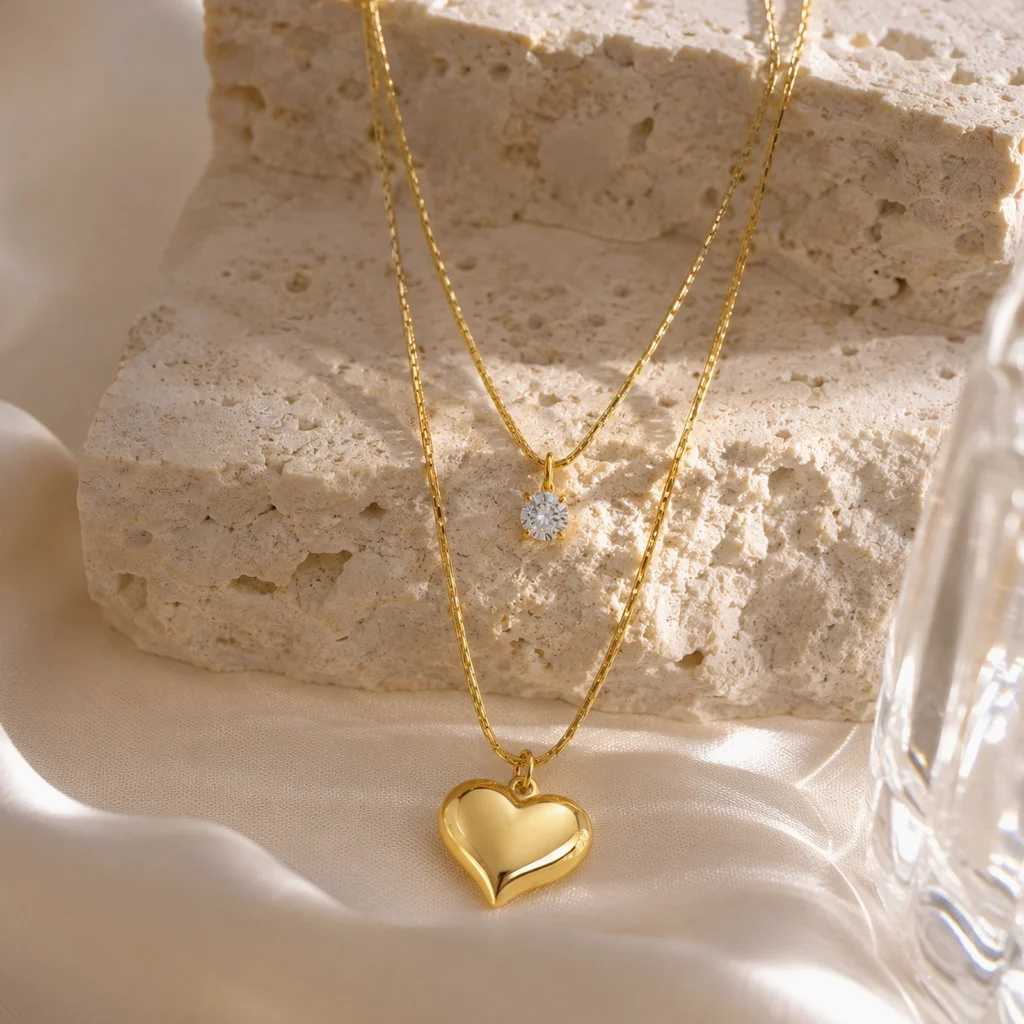 Collar Corazón Dorado Mujer chapado en Oro 18K con recubrimiento PVD sobre un escenario lifestyle premium con piedra travertino, seda y cristal de la colección Gold Essential.