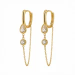 Aros Dorados Colgantes | Glow Secret Aros Dorados Colgantes de Glow Secret pertenecientes a la colección Gold Essential. Diseño dorado con aro tipo huggie, detalles de piedras transparentes y cadenas colgantes que aportan movimiento y luminosidad.