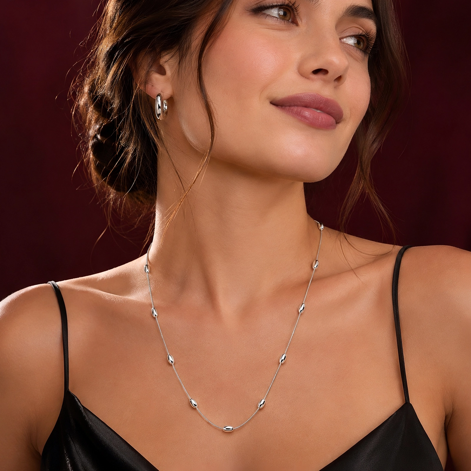 Mujer luciendo Collar Largo de Plata 925 Mujer con detalles ovalados, vestido negro y fondo burdeos de la colección ICONIC.